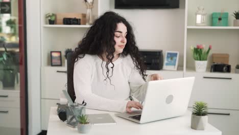 the young woman with long curly hair is sitting at a laptop in the office and working. she indicates that she's tired from work. her eyes hurt, so she puts on glasses.