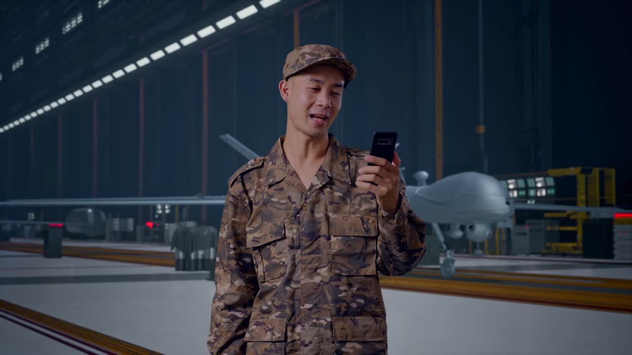Soldato asiatico che saluta con la mano e fa una videochiamata sullo smartphone mentre è in piedi con un moderno drone militare in un hangar di manutenzione.