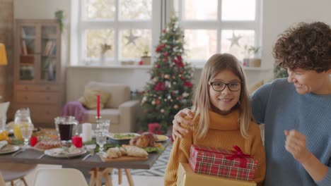 Madre-Morena-De-Pelo-Corto-Abraza-A-Su-Hija-En-La-Sala-De-Estar-Con-Mesa-Y-Adornos-Navideños