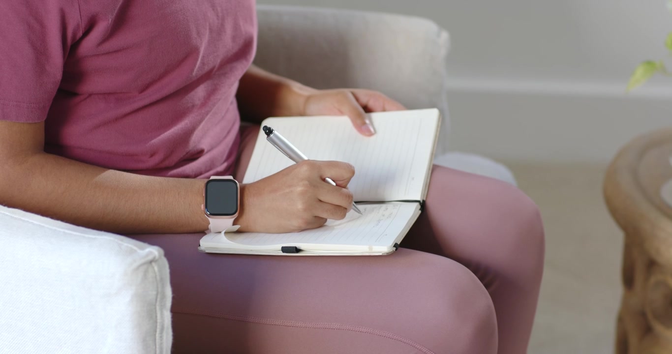 Donna seduta in poltrona, con un notebook sulle ginocchia, che scrive un diario con penna e smartwatch a casa
