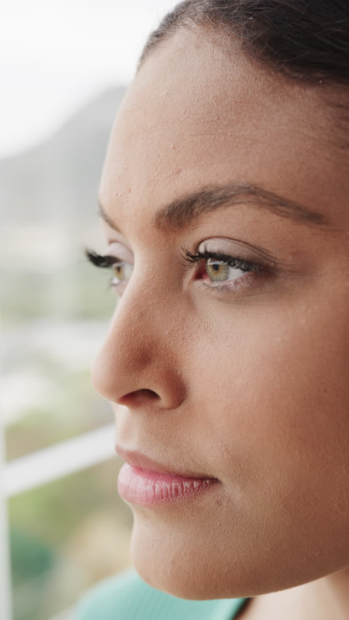 Vertical video of thoughtful biracial woman looking through window at home, slow motion