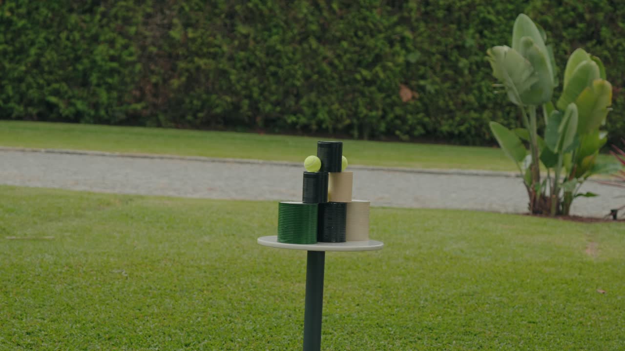 Lille rundt bord på græsset med stablede cylindriske beholdere toppet med tennisbolde i udendørs omgivelser.