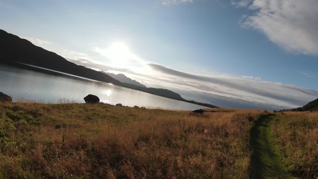 volando a lo largo de un pequeño sendero cubierto de hierba junto a un lago, girando para volar sobre el lago hacia el sol reflejado en el agua y luego de vuelta hacia el sendero con vistas a las montañas al fondo