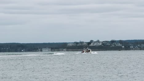 A-lobstering-or-fishing-vessel-travels-under-full-throttle-across-the-inner-harbor-with-a-backdrop-of-luxury-homes-and-a-wooded-horizon