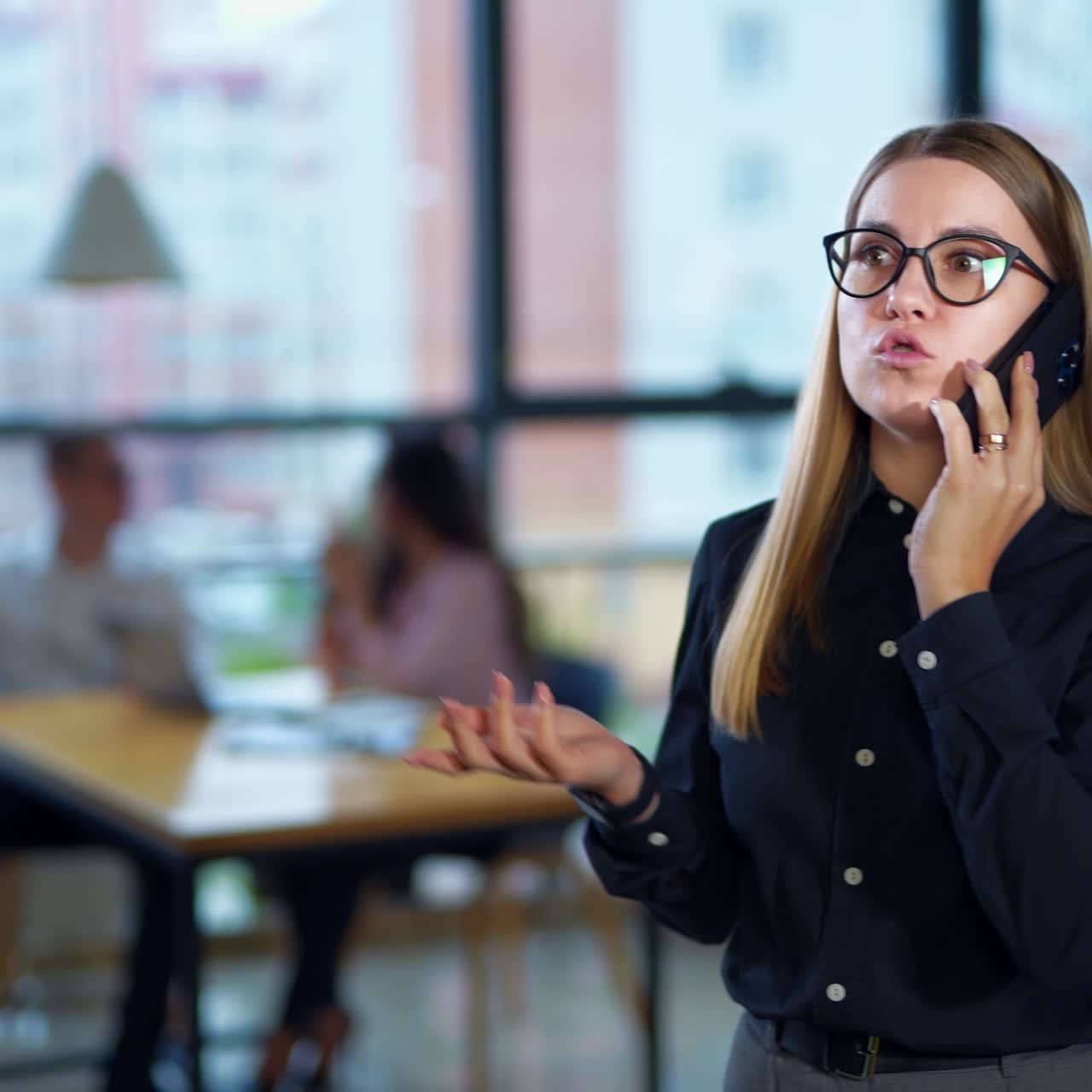 Young woman speaking on the phone and walking in the office. Emotional conversation over the phone. Couple at the table at backdrop