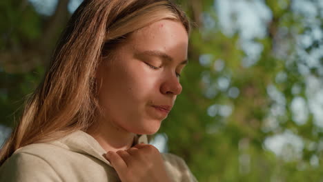 close-up of young woman with small spot on her face, deeply focused on operating her phone while holding her hood collar, sunlight filters through tree leaves, casting gentle shadows on her face