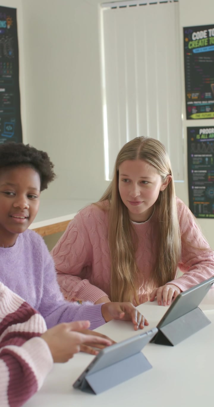 Vertical video: Diverse teen girls in class, peer tapping tablets, blonde in pink sweater learning
