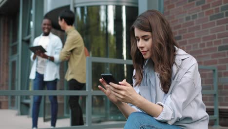 vista lateral de una estudiante caucásica usando un smartphone en la calle cerca de la universidad