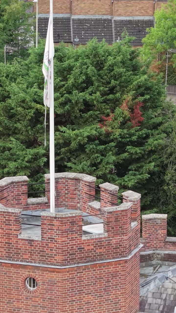 Vertical drone aerial reveal of Hertford Castle’s turret and flag, with trees, rooftops, a backdrop of greenery with a road and moving vehicles in the distance