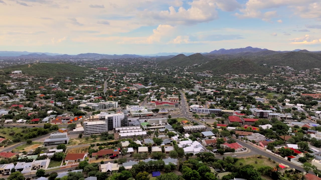 Volo aereo sui quartieri e le strade principali di Windhoek, con la città incorniciata da aspre colline. La capitale si estende in un'ampia valle circondata da aspre montagne dell'altopiano sotto un cielo aperto.