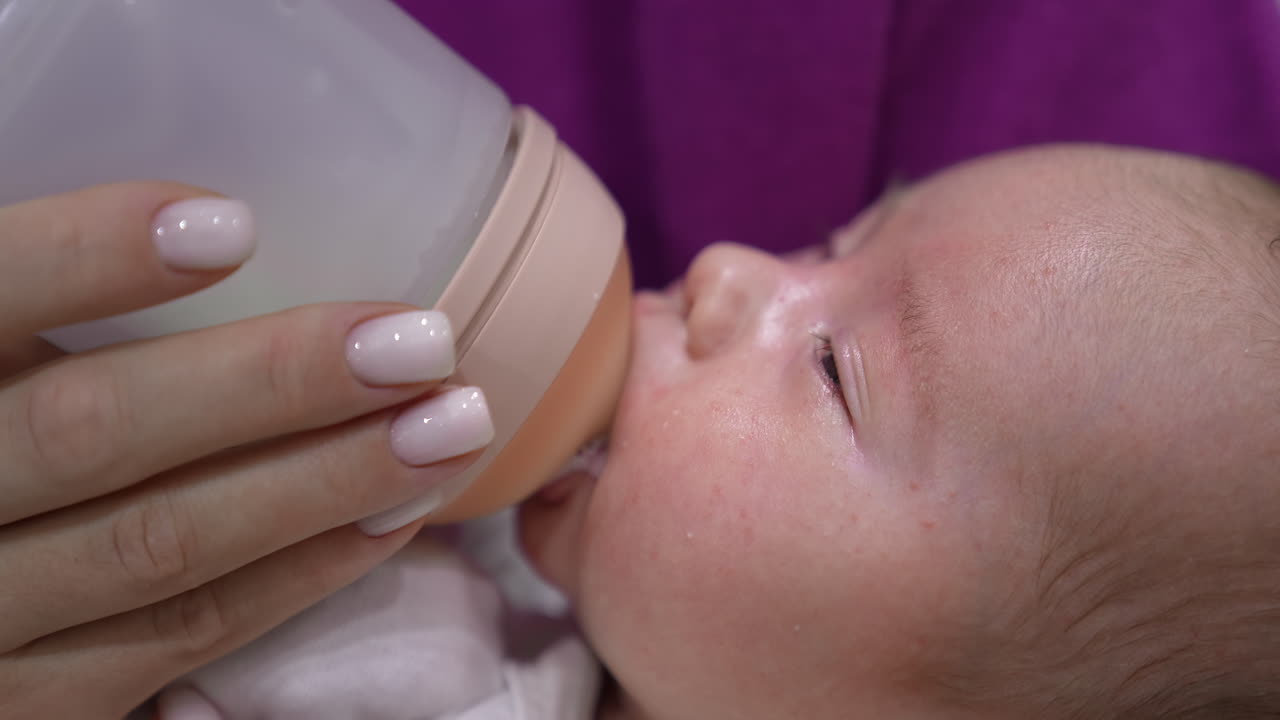 Lovely baby suckling milk from a bottle and slowly closing his eyes. Mother's hand holding a bottle for a baby. Cute baby's face close up.
