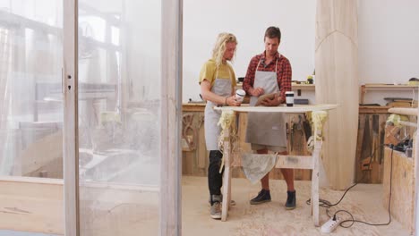 Two-Caucasian-male-surfboard-makers-standing-and-working-on-projects-using-a-tablet