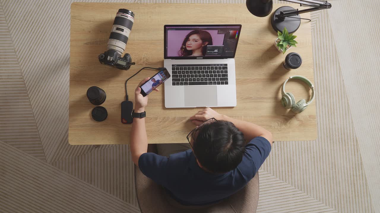 Top View Of A Male Editor Waving Hand And Having A Video Call On Smartphone While Sitting In The Workspace Using A Laptop Next To The Camera Editing Photo Of A Female At Home
