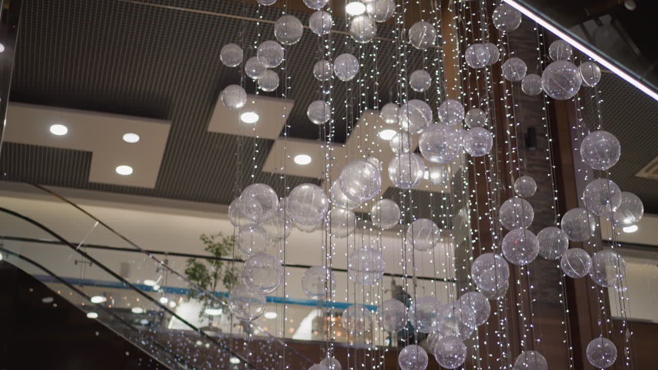 hanging transparent spheres and festive string lights draped from ceiling in mall corridor with escalator and faint silhouettes of shoppers passing by under warm decorative lighting