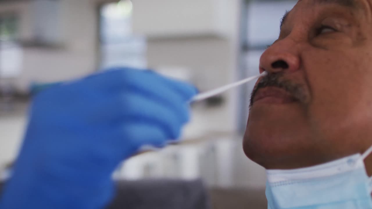 Mixed race female doctor wearing mask doing swab test on senior man at home