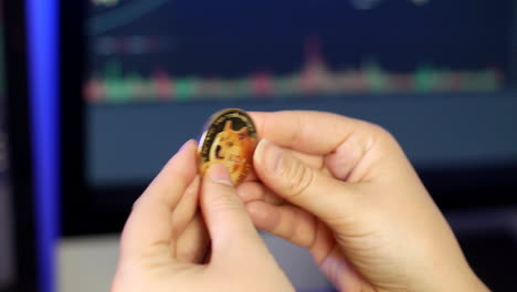 unrecognizable woman holding and looking at dogecoin in her fingers in front of cryptocurrency trading graphs