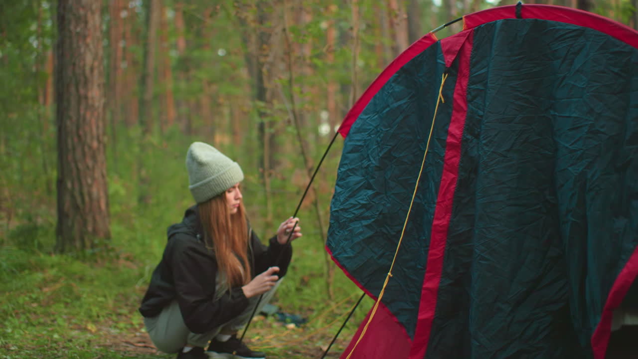 Lady crouches beside tent in forest, gripping flexible pole while setting up structure, surrounded by lush greenery and tall trees, wearing beanie and jacket