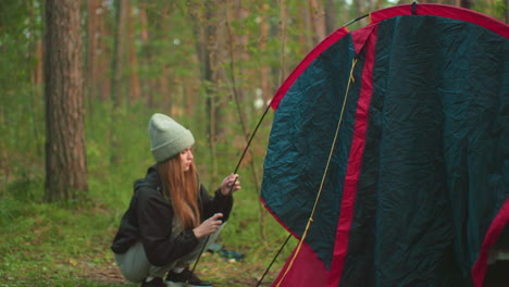 lady crouches beside tent in forest, gripping flexible pole while setting up structure, surrounded by lush greenery and tall trees, wearing beanie and jacket