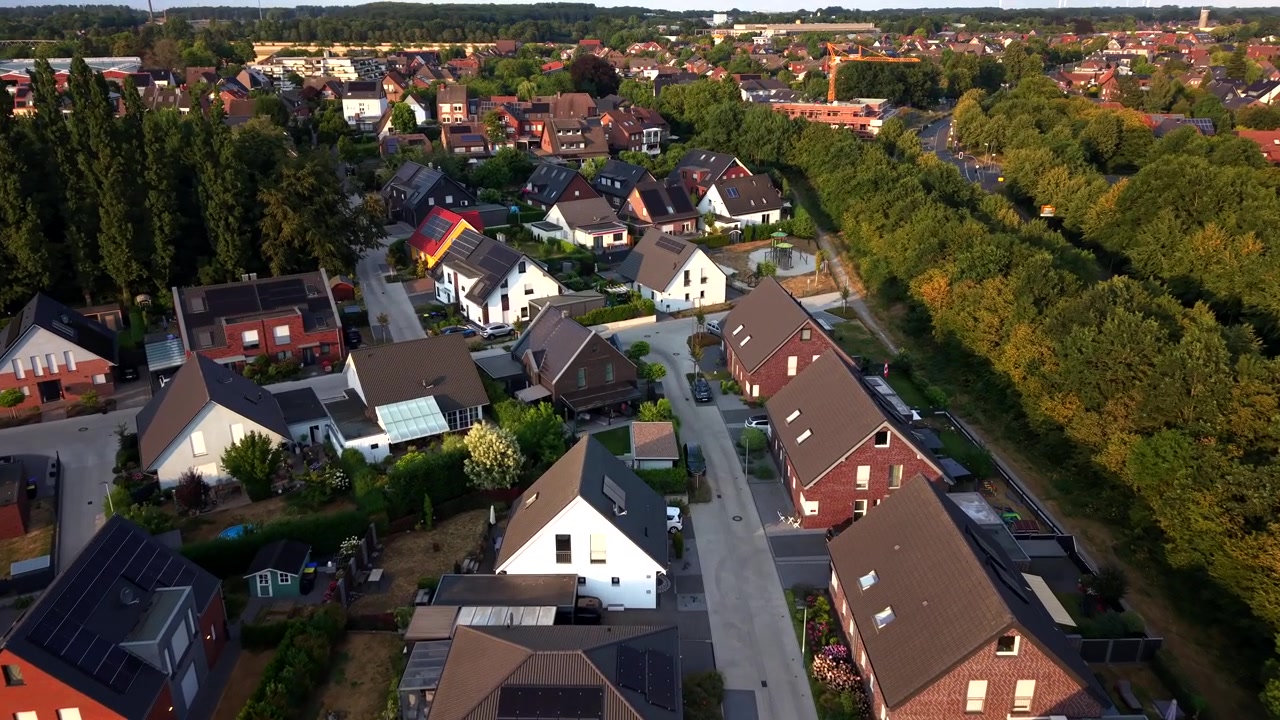 Moderne solpaneler på taget gløder under gyldent solnedgangslys over en rødtaget tysk forstad, en stille villavej snor sig mellem gavlhuse og en tæt trækant i baggrunden. Luftfoto fra drone.