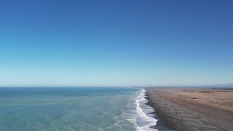 High-aerial-above-incoming-waves-from-beautiful-turquoise-ocean-with-flat,-dry-land-and-foothills-in-distance