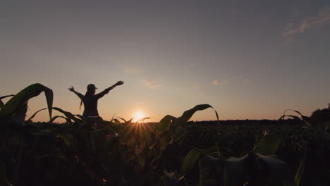 a young female farmer raises her hands over a field of corn where the sun is setting