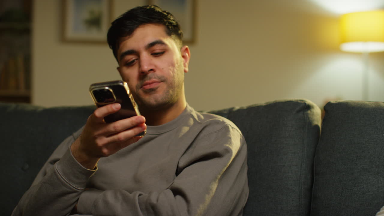 joven preocupado pasando la noche en casa sentado en el sofá con el teléfono móvil desplazándose por internet o medios sociales de transmisión de contenido