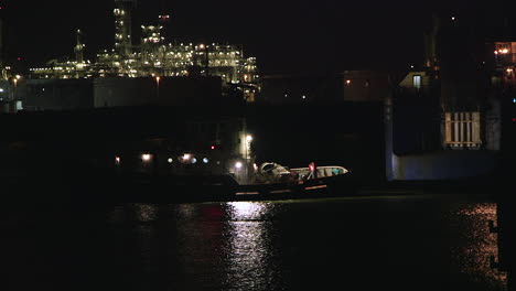 tugboat at night near an illuminated industrial refinery