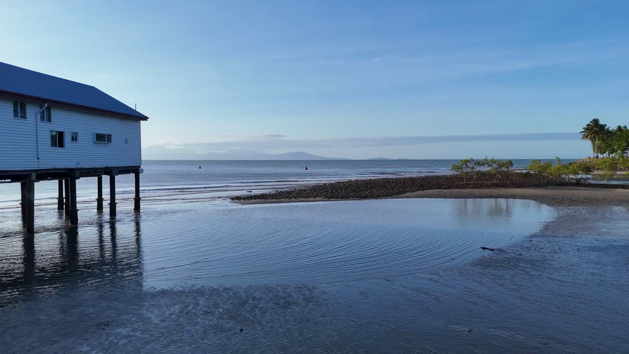 Drone captures serene coastal landscape with a pier, mangroves, and calm waters under clear skies in Port Douglas, Australia