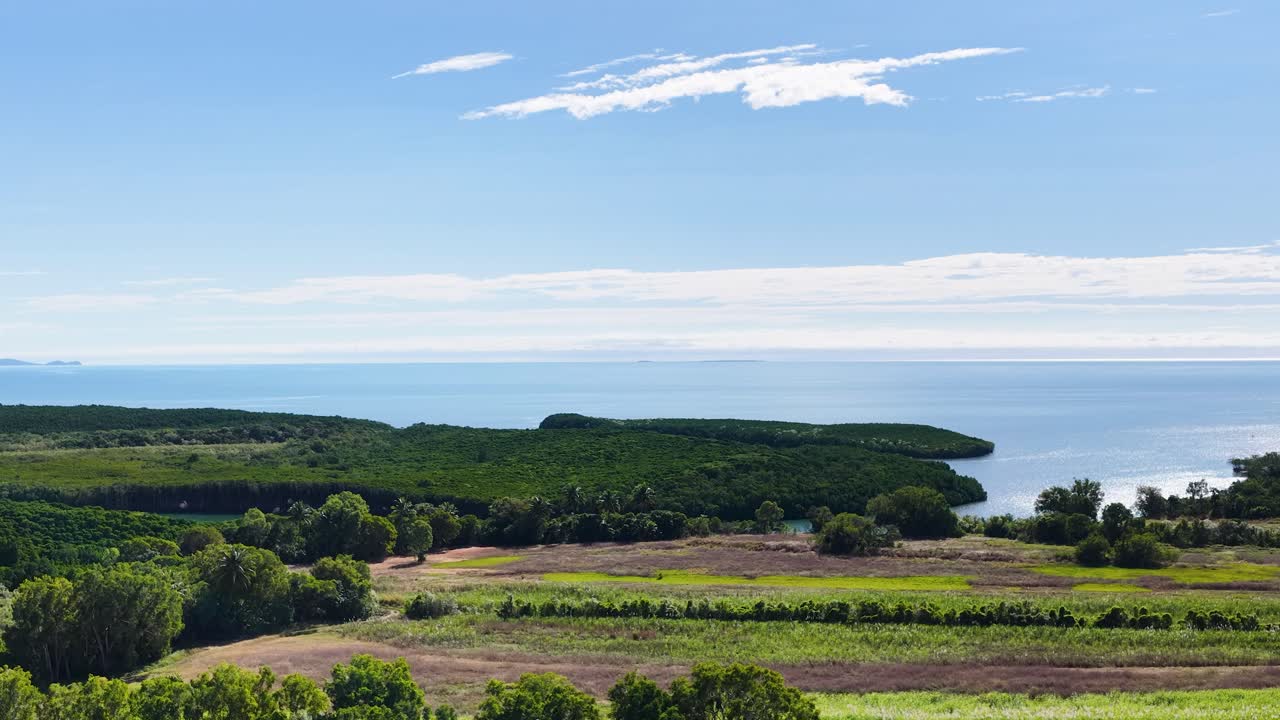 Camera slowly pans across green farmland toward distant rainforest and coastline under bright daylight