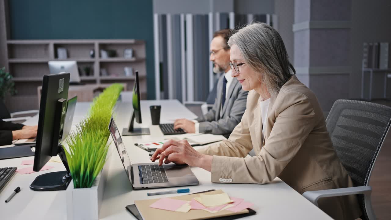 성공적 인 경험 많은 노인 여성 관리자 의복과 안경을 입고 직장에서 노트북 컴퓨터를 사용하여 추적