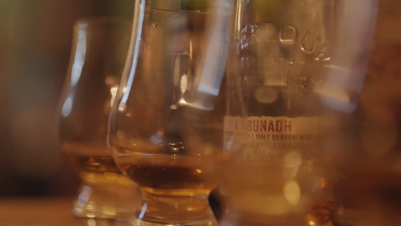 A slow, shallow-focus pan reveals several whiskey tasting glasses filled with amber liquid on a wooden bar in warm, ambient lighting