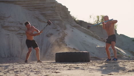 Two-powerful-male-athletes-training-together-hit-the-wheel-with-a-hammer-at-sunset-in-the-desert.-Endurance-training