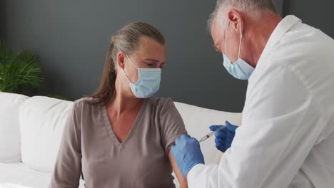 médico masculino caucásico con máscara facial dando la vacuna contra la covid-19 a una mujer en casa