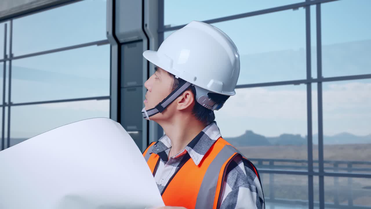 vista lateral de cerca de un ingeniero masculino asiático con casco de seguridad mirando el plano en sus manos y mirando a su alrededor mientras está de pie en una fábrica de alta tecnología, analizando el concepto de máquina robótica