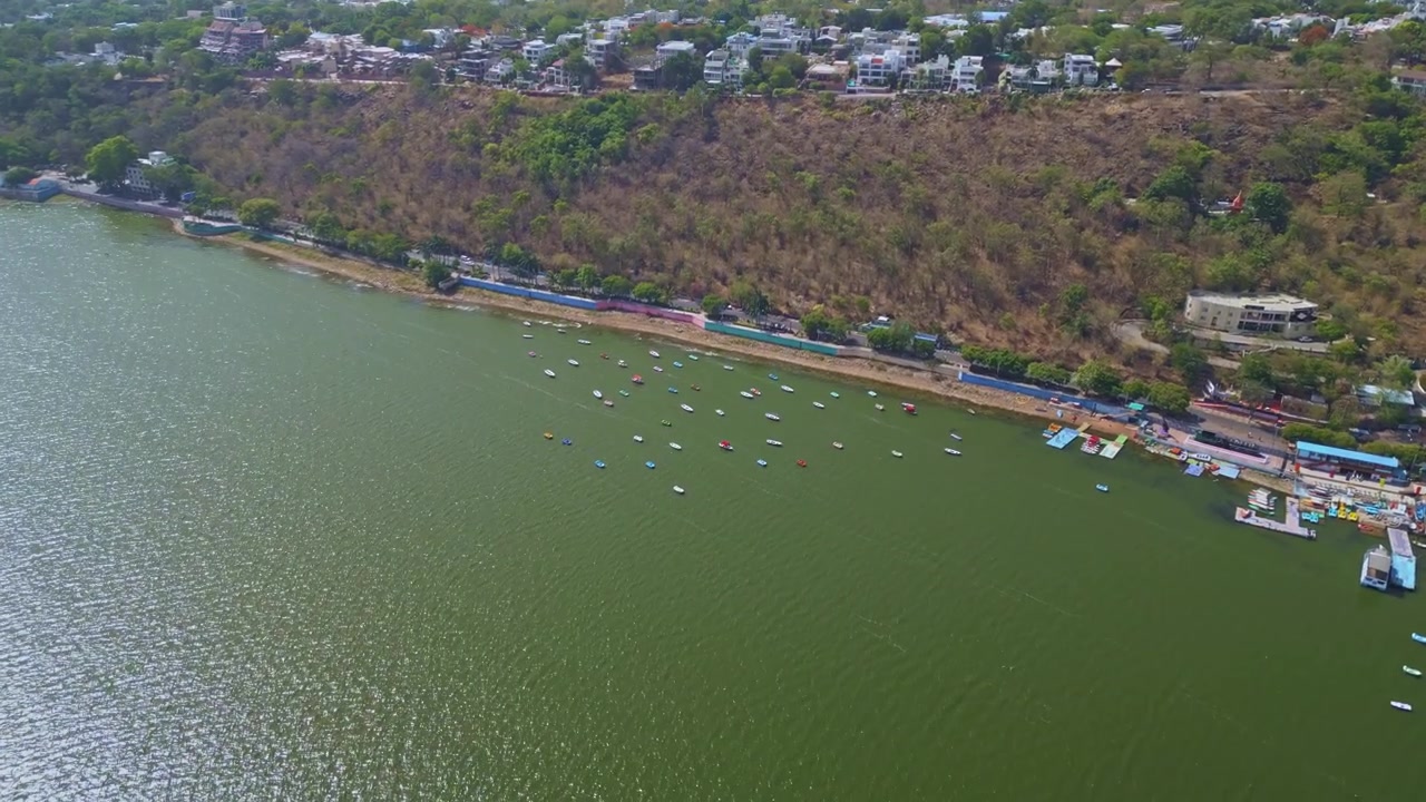 Riprese aeree con drone che avanzano attraverso il Lago Superiore verso il Boat Club Bhopal, con acque calme, barche ormeggiate, lungolago e residenze collinari nel Madhya Pradesh, India.