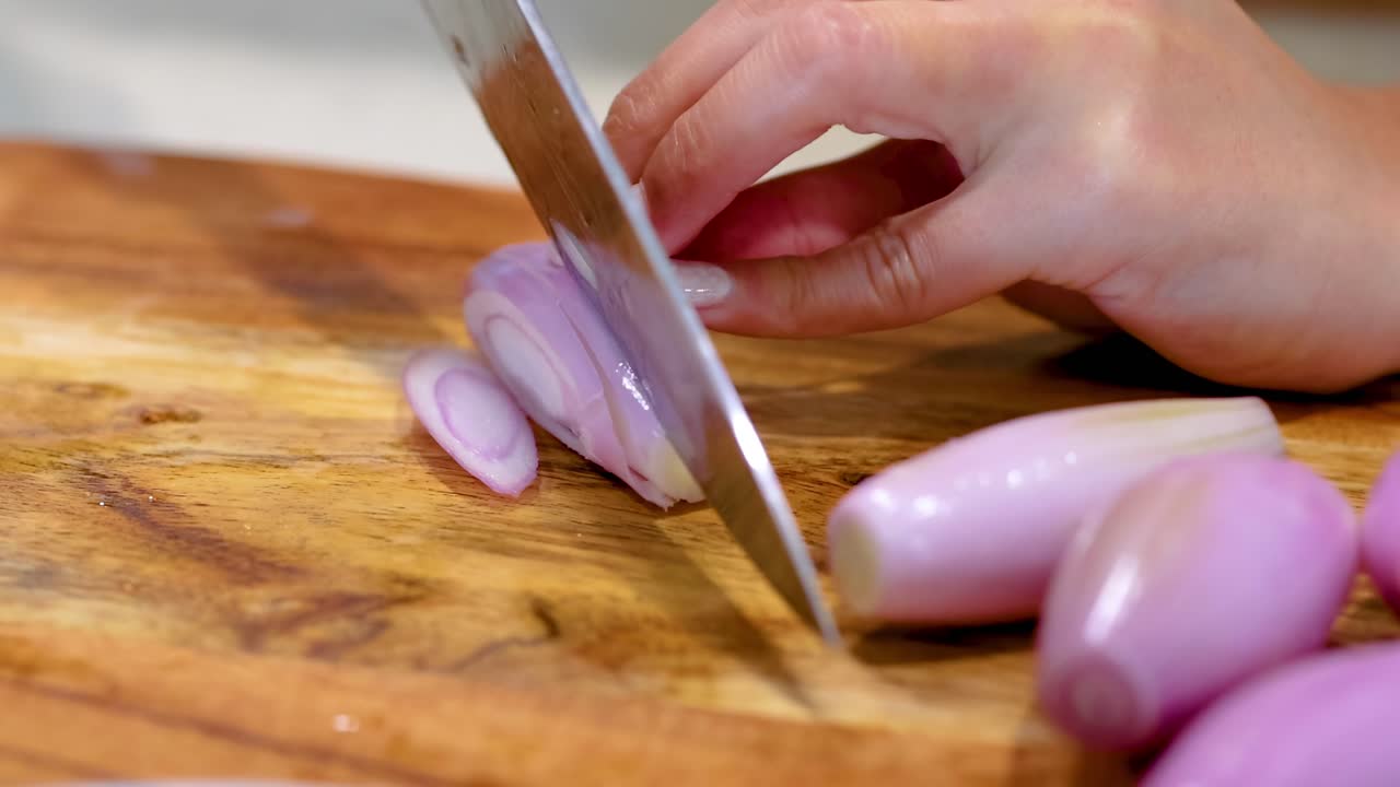 A close-up view of a hand skillfully slicing shallots on a wooden board with a sharp knife.