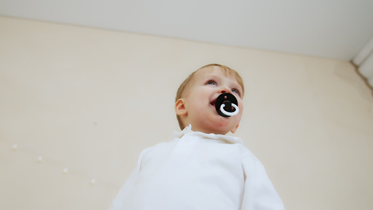 Small Caucasian toddler kid wearing white shirt holding a pacifier in mouth. Low angle view at the lovely baby boy.