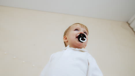 small caucasian toddler kid wearing white shirt holding a pacifier in mouth. low angle view at the lovely baby boy.