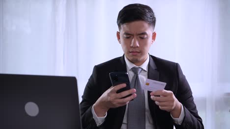 smart young asian businessman sitting at home office with laptop smiling and happy while using credit card with online shopping mobile application on smartphone.