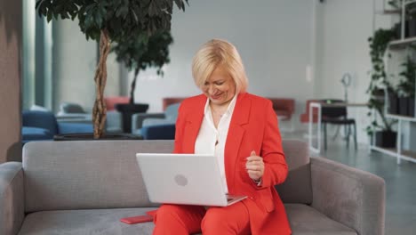 Una-Mujer-Mayor-Alegre-Con-Un-Traje-Rojo-Está-Sentada-En-Un-Sofá-En-Un-Centro-De-Negocios,-Bailando-Felizmente-Con-Una-Computadora-Portátil