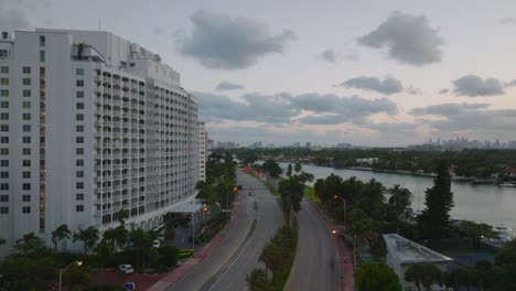 forwards fly above multilane road leading through tropical city. palm trees waving in wind at dusk. aerial view of modern borough with multistorey buildings. miami, usa