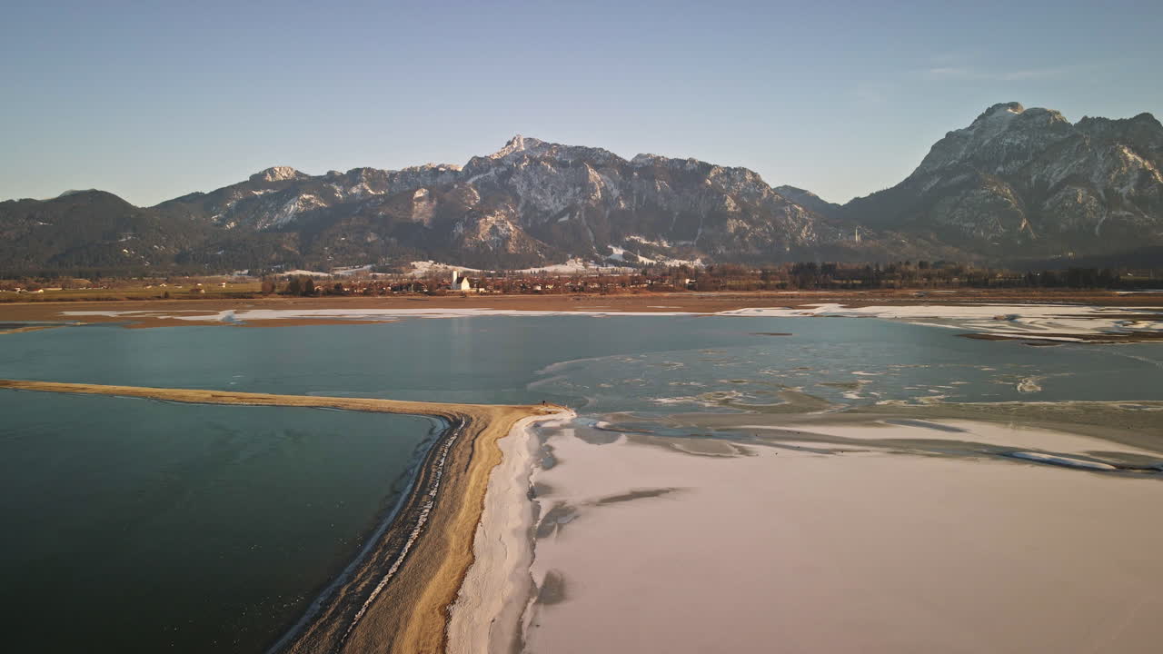 Camera glides forward along a narrow sandbar where snow and ice meet deep blue water, with Bavarian mountains and a winter village in the distance