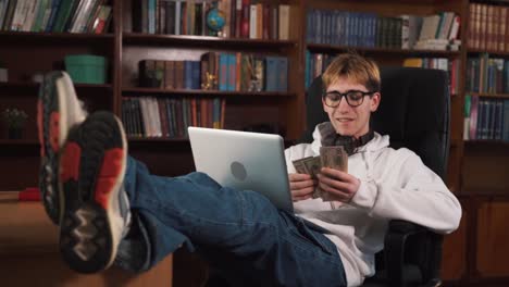 The-handsome-guy-is-sitting-in-a-chair,-with-his-feet-on-the-table,-and-he-holds-a-laptop-on-his-lap