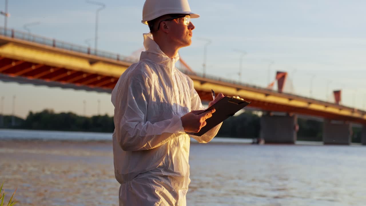 Inspector in protective clothing stands by waterfront reviewing contamination risks in warm daylight