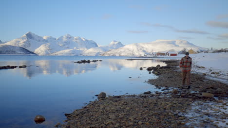 A-Man-Walking-Along-a-Tranquil-Lake-with-the-Reflection-of-Mountains-in-an-Amazing-Winter-Environment,-Static-Slow-Motion