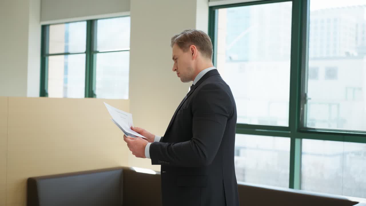 A thoughtful businessman carefully reviewing documents in his hands standing by a window in a bright modern office space while natural light streams through green-framed windows behind him - orbit