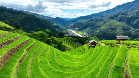 drone image of layered rice paddies stretching across hilly terrain, featuring small wooden shelters, vibrant fields, and a peaceful countryside village nestled among forested slopes.