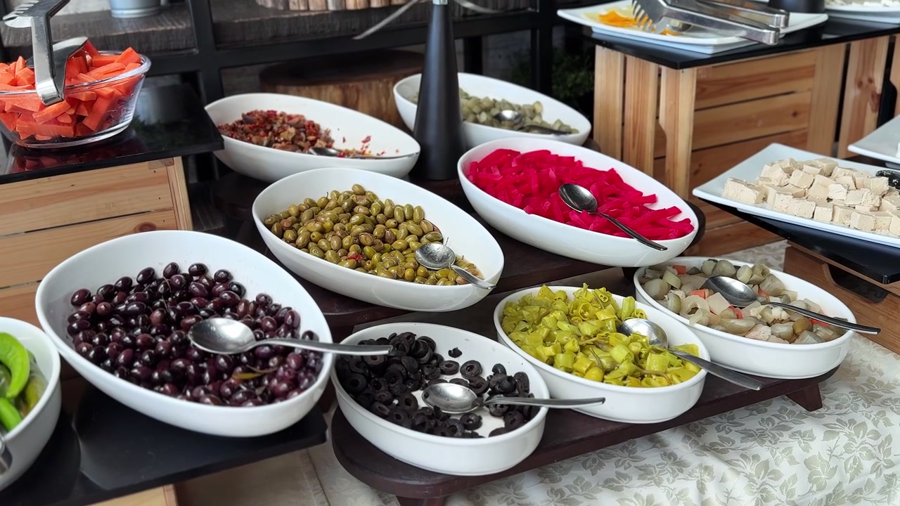 Una vista ravvicinata di una variegata stazione buffet di catering al coperto durante il giorno. Ciotole di ceramica bianca mostrano un assortimento di olive fresche nere e verdi, verdure sottaceto e carote a fette