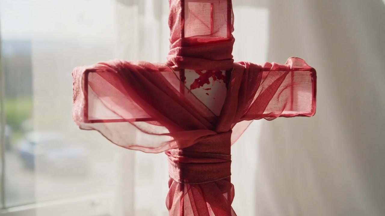 Standing wrapped wooden cross catching sunlight near window, causing red scarf lifting and swaying