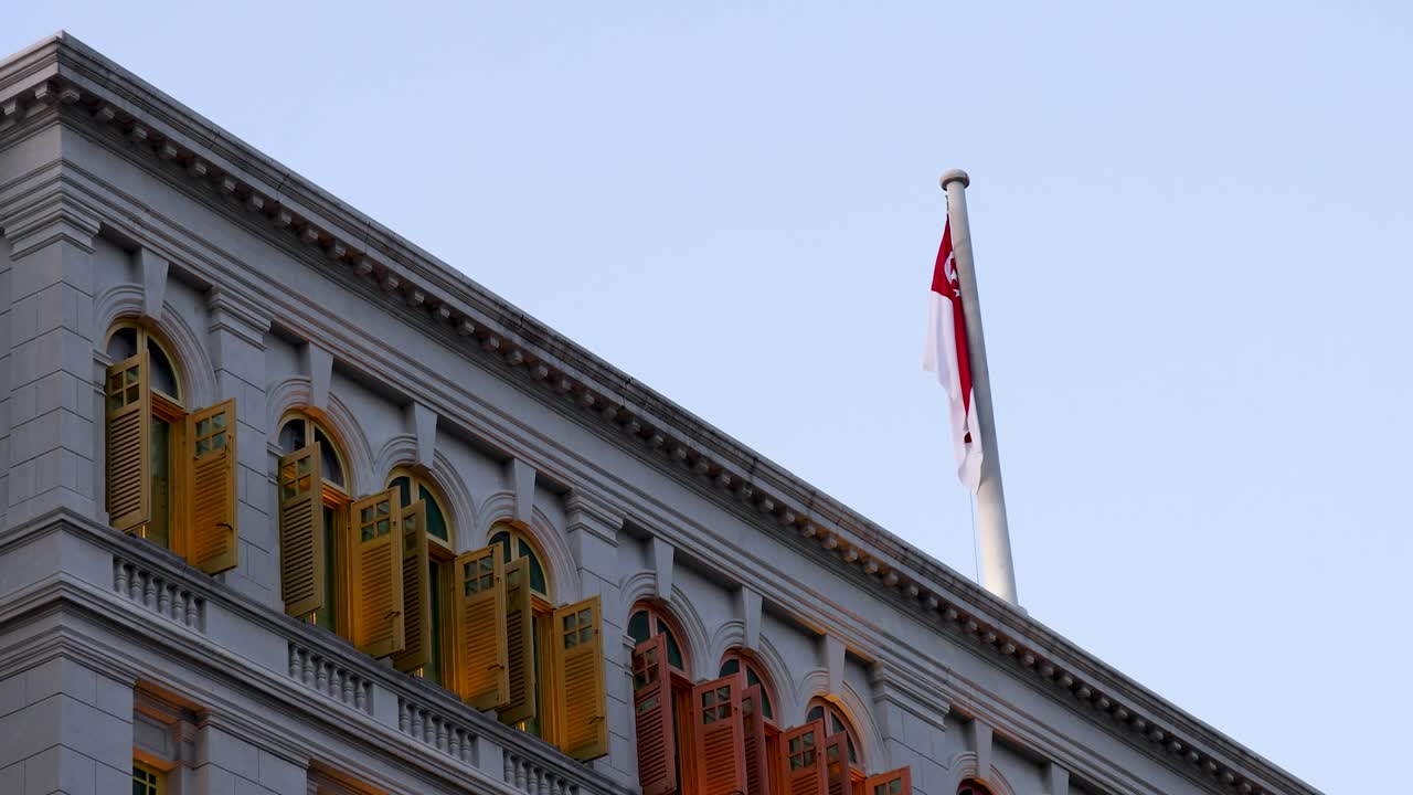 Singapore flag waves atop ornate building with colorful window shutters, captured in steady daylight shot